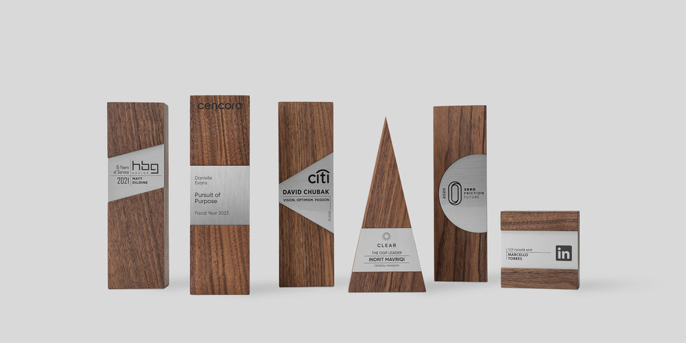 A collection of six unique corporate awards made from walnut wood with brushed metal accents. Each award is uniquely customized and engraved with company logos and recipient names, showcasing a variety of meaningful employee recognition.
